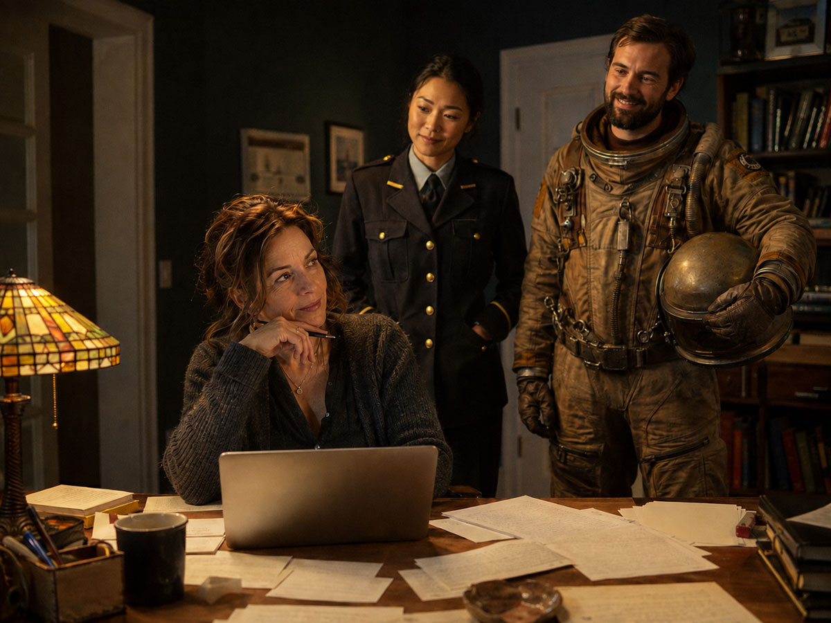 A novelist writing at her desk at night, with two characters from her novel standing easily behind her: a woman in a station officer's uniform and a mine worker in a scuffed pressure suit holding his helmet